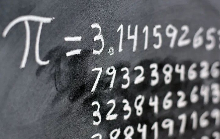Na zdjęciu widać fragment tablicy lub ciemnego tła, na którym białą kredą zapisano symbol matematyczny liczby π (pi) oraz jej rozwinięcie dziesiętne zaczynające się od 3,141592653…. Liczby zapisane są w kilku liniach, a część zapisu jest lekko rozmazana, jakby ktoś próbował zetrzeć fragment kredy. Obraz przedstawia motyw matematyczny związany z liczbą pi