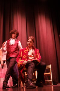 Dwóch aktorów na scenie w czerwonym oświetleniu. Jedna osoba siedzi na krześle w ozdobnym, czerwonym stroju, druga stoi obok i gestykuluje. W tle widoczna kurtyna i fragmenty scenografii.