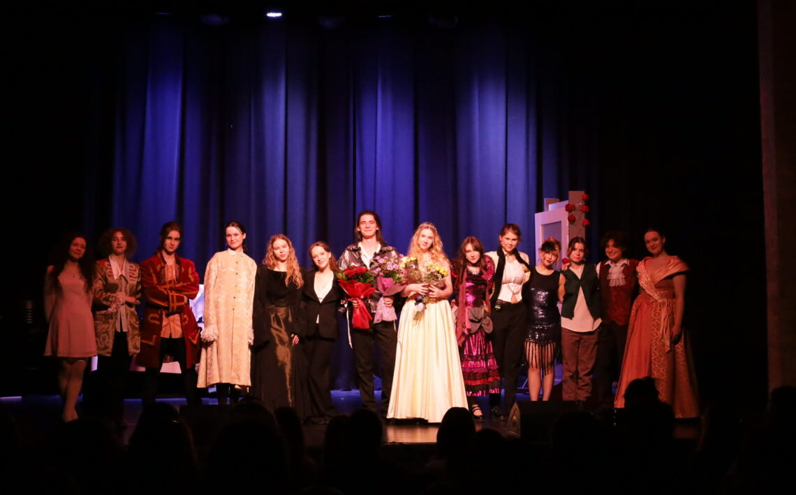 Grupa kilkunastu młodych aktorów stoi w rzędzie na scenie przed kurtyną. W centrum dwie osoby trzymają bukiety kwiatów. Wszyscy ubrani w różnorodne kostiumy teatralne, widoczna publiczność w dolnej części kadru.