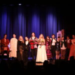 Grupa kilkunastu młodych aktorów stoi w rzędzie na scenie przed kurtyną. W centrum dwie osoby trzymają bukiety kwiatów. Wszyscy ubrani w różnorodne kostiumy teatralne, widoczna publiczność w dolnej części kadru.