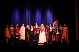 Grupa kilkunastu młodych aktorów stoi w rzędzie na scenie przed kurtyną. W centrum dwie osoby trzymają bukiety kwiatów. Wszyscy ubrani w różnorodne kostiumy teatralne, widoczna publiczność w dolnej części kadru.