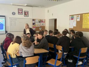 W sali lekcyjnej uczniowie siedzą w kilku grupach przy połączonych ławkach i pracują razem nad zadaniem. Nauczycielka chodzi między stolikami i pomaga uczniom. Na ścianach widać tablice z ogłoszeniami, kalendarz oraz krzyż.