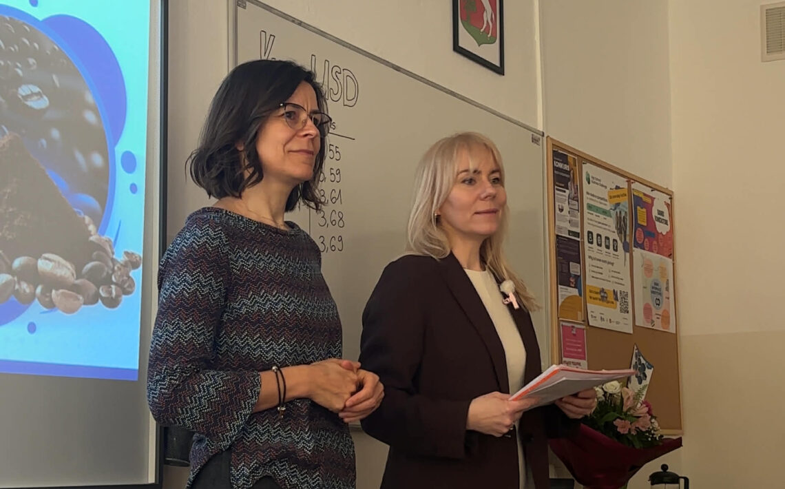 Dwie nauczycielki stoją z przodu klasy obok tablicy i ekranu z prezentacją. Jedna z nich trzyma notatki. Za nimi na tablicy zapisany jest temat „Kurs USD” z kilkoma wartościami liczbowymi. Na ścianie wiszą godła oraz tablica z ogłoszeniami.