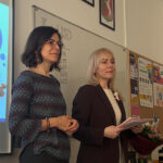 Dwie nauczycielki stoją z przodu klasy obok tablicy i ekranu z prezentacją. Jedna z nich trzyma notatki. Za nimi na tablicy zapisany jest temat „Kurs USD” z kilkoma wartościami liczbowymi. Na ścianie wiszą godła oraz tablica z ogłoszeniami.