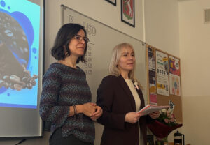 Dwie nauczycielki stoją z przodu klasy obok tablicy i ekranu z prezentacją. Jedna z nich trzyma notatki. Za nimi na tablicy zapisany jest temat „Kurs USD” z kilkoma wartościami liczbowymi. Na ścianie wiszą godła oraz tablica z ogłoszeniami.