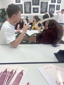 Uczniowie w klasie korzystają z gogli VR; jedna osoba instruuje drugą, inni w tle również testują sprzęt.