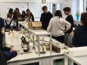 Pracownia chemiczna. Uczniowie stoją wokół stołów laboratoryjnych, wykonują doświadczenia. Na stołach znajdują się probówki, odczynniki chemiczne i sprzęt laboratoryjny.