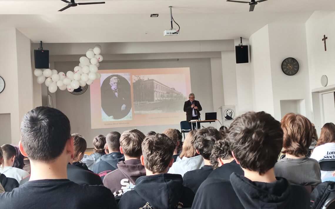 Widok sali szkolnej lub auli z perspektywy siedzącej publiczności. Na pierwszym planie widać tyły głów kilkunastu uczniów siedzących w rzędach na krzesłach. Przed nimi, na środku, stoi starszy mężczyzna (siwe włosy, garnitur), który przemawia, trzymając mikrofon. Obok niego znajduje się mały stolik z laptopem oraz krzesło. Za nim na ścianie wyświetlany jest slajd z projektora – po lewej portret starszego mężczyzny, po prawej zdjęcie dużego budynku (prawdopodobnie szkoły). Nad sceną wiszą dwa czarne głośniki i projektor sufitowy. Po lewej stronie dekoracja z białych balonów. Po prawej ścianie widoczny zegar oraz krzyż. Pomieszczenie jest jasne, o białych ścianach i drewnianym suficie.