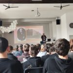 Widok sali szkolnej lub auli z perspektywy siedzącej publiczności. Na pierwszym planie widać tyły głów kilkunastu uczniów siedzących w rzędach na krzesłach. Przed nimi, na środku, stoi starszy mężczyzna (siwe włosy, garnitur), który przemawia, trzymając mikrofon. Obok niego znajduje się mały stolik z laptopem oraz krzesło. Za nim na ścianie wyświetlany jest slajd z projektora – po lewej portret starszego mężczyzny, po prawej zdjęcie dużego budynku (prawdopodobnie szkoły). Nad sceną wiszą dwa czarne głośniki i projektor sufitowy. Po lewej stronie dekoracja z białych balonów. Po prawej ścianie widoczny zegar oraz krzyż. Pomieszczenie jest jasne, o białych ścianach i drewnianym suficie.