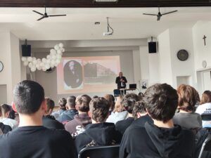 Widok sali szkolnej lub auli z perspektywy siedzącej publiczności. Na pierwszym planie widać tyły głów kilkunastu uczniów siedzących w rzędach na krzesłach. Przed nimi, na środku, stoi starszy mężczyzna (siwe włosy, garnitur), który przemawia, trzymając mikrofon. Obok niego znajduje się mały stolik z laptopem oraz krzesło. Za nim na ścianie wyświetlany jest slajd z projektora – po lewej portret starszego mężczyzny, po prawej zdjęcie dużego budynku (prawdopodobnie szkoły). Nad sceną wiszą dwa czarne głośniki i projektor sufitowy. Po lewej stronie dekoracja z białych balonów. Po prawej ścianie widoczny zegar oraz krzyż. Pomieszczenie jest jasne, o białych ścianach i drewnianym suficie.