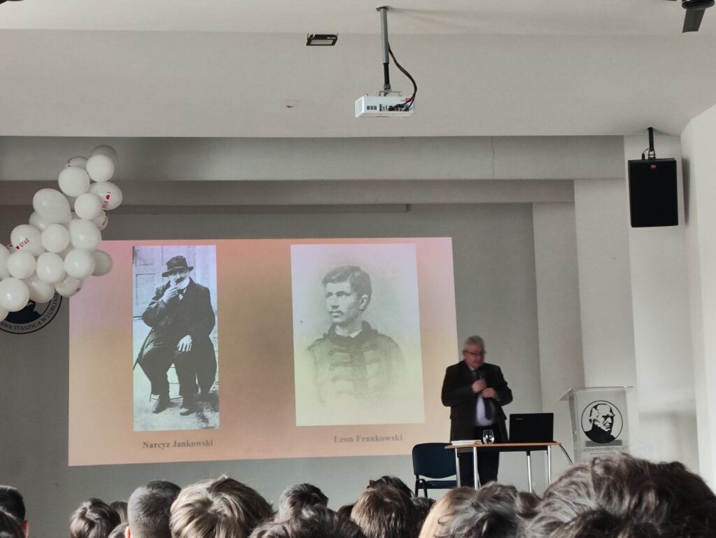 Zbliżony widok tej samej sceny. Na środku stoi ten sam mężczyzna w garniturze, przemawiający do uczniów. Na ekranie za nim wyświetlone są dwa czarno-białe portrety mężczyzn podpisane: „Narcyz Jankowski” oraz „Leon Frankowski”. Na pierwszym zdjęciu mężczyzna siedzi w kapeluszu, na drugim młodszy mężczyzna w formalnym stroju. W dolnej części kadru widoczne są głowy uczniów siedzących na widowni. Nad ekranem znajduje się projektor, a po bokach ściany wiszą czarne głośniki. Po lewej stronie częściowo widoczna dekoracja z białych balonów. Pomieszczenie jest dobrze oświetlone i uporządkowane.