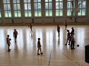 Sala gimnastyczna z boiskiem do koszykówki. Kilku uczniów w strojach sportowych stoi i trenuje z piłkami. W tle duże okna i widok na boisko na zewnątrz.