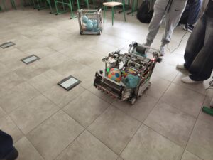 Na podłodze sali stoi robot zbudowany z metalowej konstrukcji, przewożący kolorowe piłki. Wokół stoją uczniowie (widoczne nogi), obserwują działanie urządzenia.