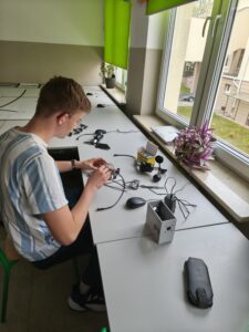 Uczeń siedzi przy stanowisku komputerowym przy oknie. Montuje układ elektroniczny z przewodów i płytki. Na biurku leżą narzędzia, kamera, myszka i elementy techniczne, obok roślina doniczkowa.