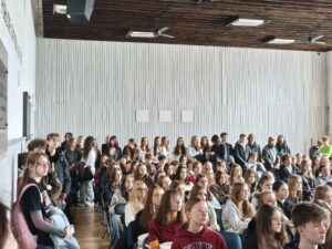 Duża grupa uczniów siedzi i stoi w przestronnej sali szkolnej. Większość patrzy w jednym kierunku, jakby słuchali wystąpienia. Wysokie ściany, drewniany sufit i jasne oświetlenie dzienne.