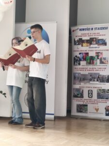 Dwoje uczniów (nastolatków) stoi na scenie lub sali szkolnej. Chłopak w białej koszulce trzyma mikrofon i czyta z czerwonej teczki, obok dziewczyna również czyta z podobnej teczki. W tle widoczny roll-up z logo szkoły oraz drugi plakat z informacjami (m.in. „Nowości w Staszicu”, radio uczniowskie, zajęcia dodatkowe).