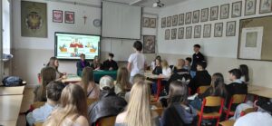 Uczniowie siedzą w klasie ustawionej jak sala sądowa; jedna osoba stoi i przemawia, a inni odgrywają role (sędziowie, świadkowie, strony), przed nimi ekran z diagramem „Sala rozpraw”.