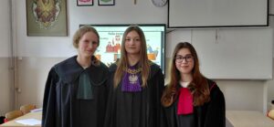 Trzy uczennice w togach stoją razem w klasie, pozując do zdjęcia na tle ekranu z prezentacją o sali rozpraw.