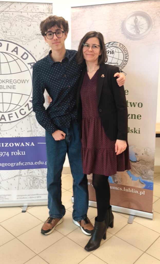 Młody chłopak i dorosła kobieta stoją razem, obejmując się, na tle roll-upów związanych z olimpiadą geograficzną; oboje patrzą w stronę aparatu i lekko się uśmiechają.