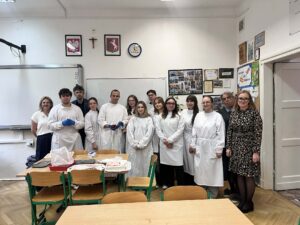 Grupa uczniów i nauczycieli stoi w klasie biologicznej; uczniowie w białych fartuchach po zajęciach sekcyjnych, na stole przed nimi znajdują się tace i materiały do pracy.