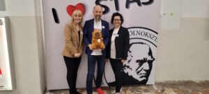 Trzy osoby stoją przed ścianką z grafiką i napisem szkoły. Mężczyzna trzyma pluszowego misia, a dwie kobiety stoją po bokach.