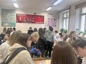 Klasa pełna uczniów pracujących w grupach. Na ścianie widoczne plakaty edukacyjne, w tym duży czerwony baner „Masz serce? Daj serce!”. Uczniowie rozmawiają i wykonują zadania.