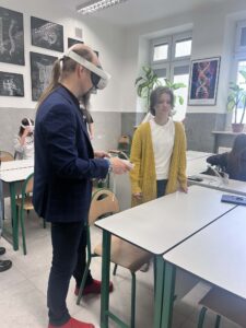 W klasie nauczyciel (mężczyzna) oraz uczennica stoją przy ławkach. Oboje korzystają z technologii VR (nauczyciel w goglach). W tle inni uczniowie również testują sprzęt.