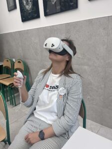 Kobieta siedzi na krześle i korzysta z gogli VR, trzyma kontroler w dłoni. Ma na sobie jasny strój z paskami. W tle krzesła i ściana z grafikami anatomicznymi.