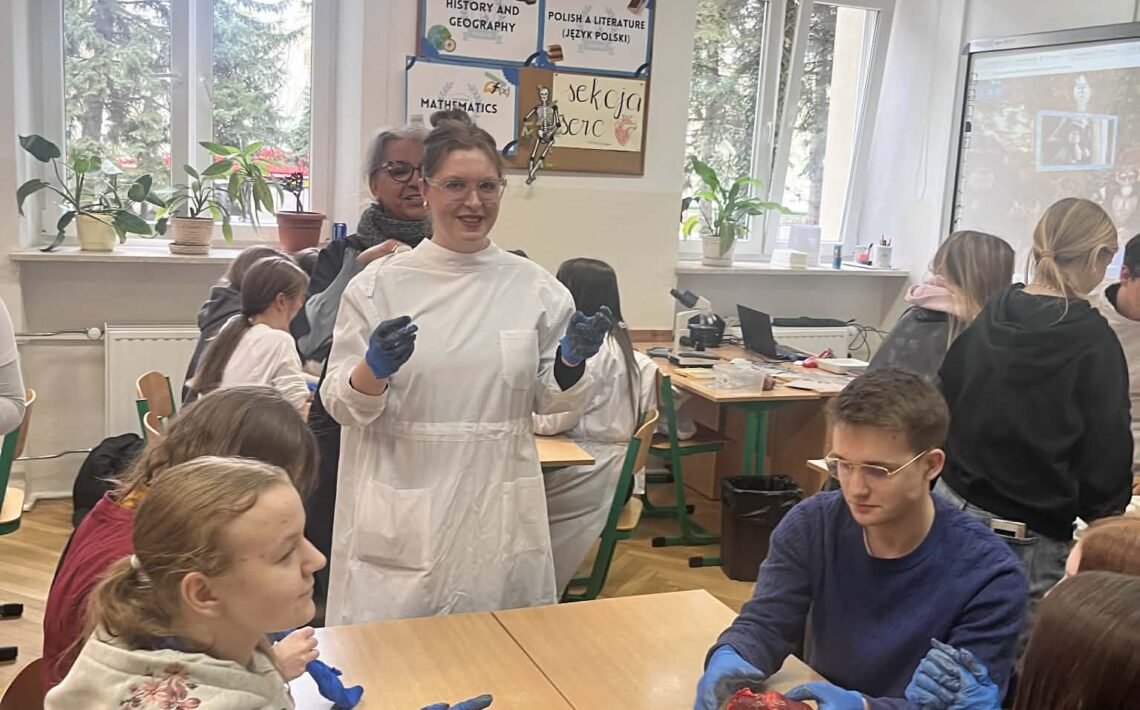 Sala lekcyjna. Uczniowie w rękawiczkach ochronnych uczestniczą w zajęciach biologicznych – na stole leży preparat (serce), które analizują. Nauczycielka stoi i tłumaczy.