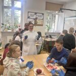 Sala lekcyjna. Uczniowie w rękawiczkach ochronnych uczestniczą w zajęciach biologicznych – na stole leży preparat (serce), które analizują. Nauczycielka stoi i tłumaczy.