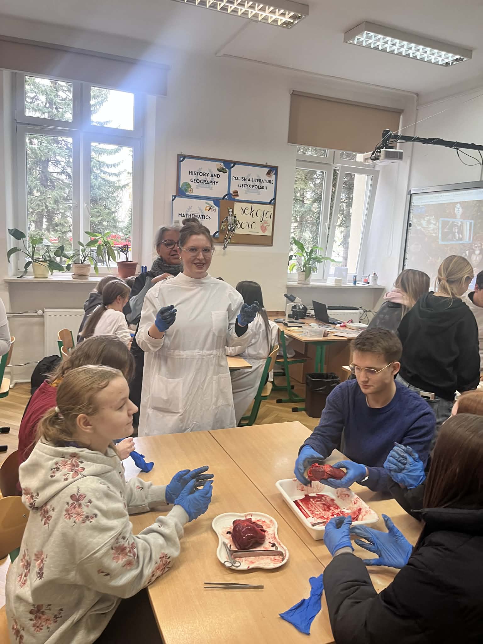 Sala lekcyjna. Uczniowie w rękawiczkach ochronnych uczestniczą w zajęciach biologicznych – na stole leży preparat (serce), które analizują. Nauczycielka stoi i tłumaczy.
