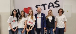 Sześć młodych osób i jeden dorosły mężczyzna stoją w rzędzie na tle dużego napisu „I ♥ STAŚ”. Jedna osoba trzyma tabliczkę „Staszic tour”. Wszyscy ubrani głównie na biało i w dżinsy.
