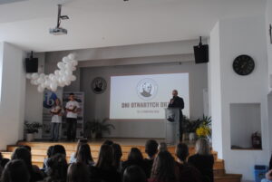 Uroczystość szkolna na sali – prowadzący przy mównicy i prezentacja „Dni Otwartych Drzwi” przed publicznością.