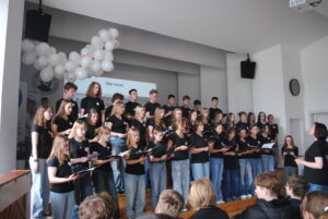Chór uczniów ubranych na czarno śpiewa na scenie, trzymając nuty w rękach.