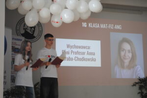 Dwójka uczniów prezentuje na scenie klasę i wychowawcę, czytając z kartek do mikrofonu.