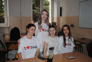 Cztery uczennice w koszulkach „I ♥ Staś” siedzą i stoją przy ławce, uśmiechają się do zdjęcia.