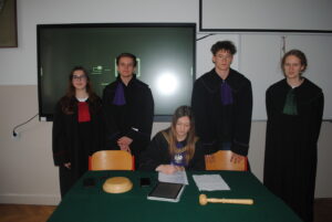 Grupa uczniów w togach stoi za stołem sędziowskim, a jedna osoba siedzi i podpisuje dokument; na stole leży młotek sędziowski.