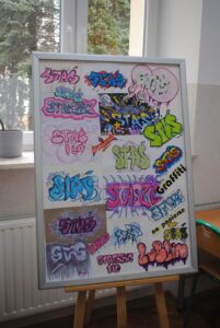 Tablica z kolorowymi pracami graficznymi przedstawiającymi napisy „Staś” w stylu graffiti.