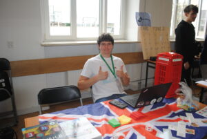 Uczeń siedzi przy stanowisku z flagą Wielkiej Brytanii i laptopem, pokazuje gest „ok” obiema rękami.