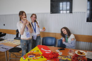 Trzy uczennice stoją przy stole z materiałami (m.in. flaga Hiszpanii), dwie trzymają jedzenie i uśmiechają się.