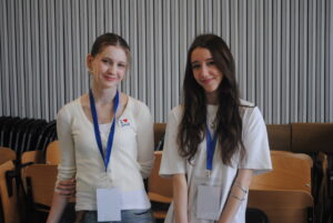 Dwie uczennice stoją obok siebie, uśmiechają się do zdjęcia i mają identyfikatory na szyi.