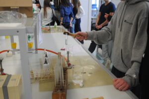 Uczeń w pracowni chemicznej trzyma probówkę nad palnikiem; na stole widoczne szkło laboratoryjne i odczynniki.