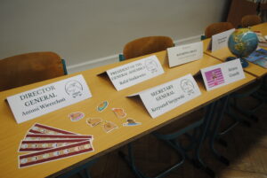 Rząd ławek z kartkami identyfikacyjnymi (np. „Director General”, „Secretary General”), małą globusem i naklejkami.