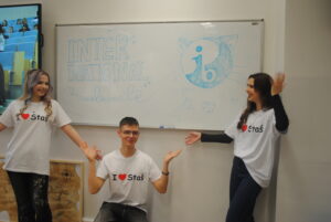 Trzy osoby w koszulkach „I ♥ Staś” stoją przy tablicy z napisem „International Baccalaureate”, prezentując program gestami.