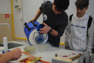 Dwóch uczniów wykonuje doświadczenie – jeden przelewa ciekły azot z pojemnika, tworząc parę, drugi obserwuje.