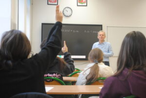 Uczniowie siedzą w ławkach, a część z nich podnosi ręce do odpowiedzi. Nauczyciel stoi przed tablicą i prowadzi lekcję.