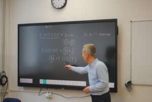 Nauczyciel matematyki stoi przy dużej tablicy multimedialnej i wskazuje zapisane równanie z wieloma cyframi „5” oraz oznaczeniami liczb pierwszych.