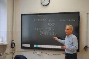 Nauczyciel przy tablicy multimedialnej tłumaczy zadanie matematyczne, wskazując zapis markerem.