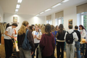 Grupa uczniów stoi i rozmawia w klasie podczas wydarzenia, w tle stoliki z materiałami i opiekunowie.