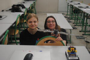 Dwie uczennice siedzą w sali komputerowej i pokazują małego drona oraz robota z klocków na stole.